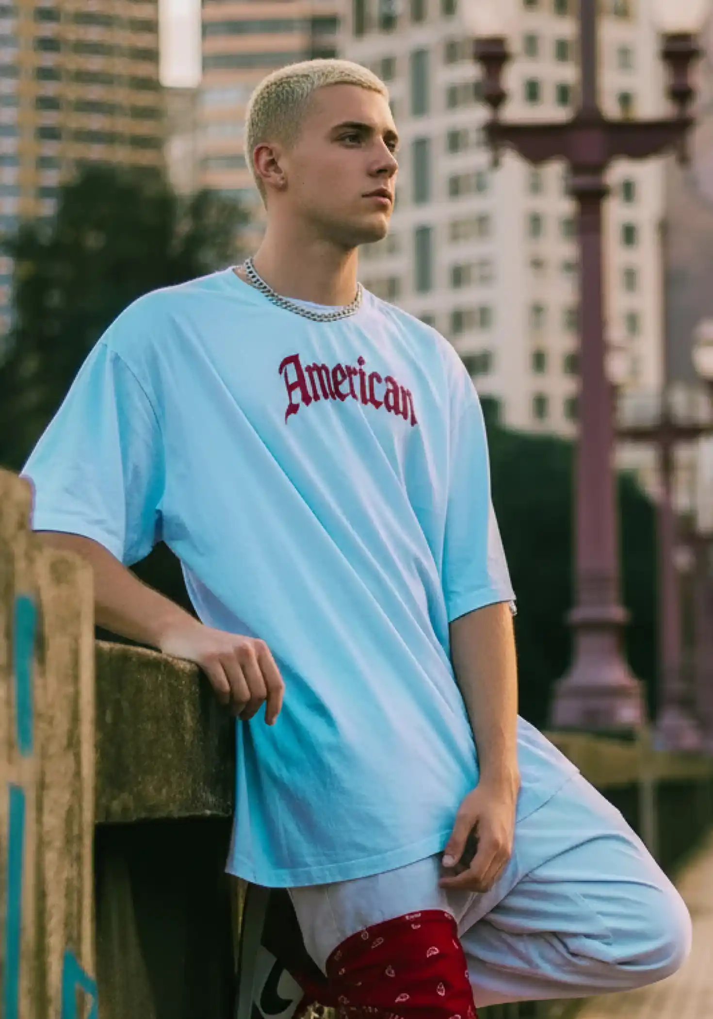 A man, wearing a custom T-shirt made by Tapstitch, is leaning against the wall. In the middle of the light blue shirt, there are red letters saying "Amertican"