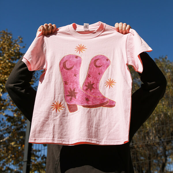 Pink graphic t-shirt with cowboy boots print held outdoors