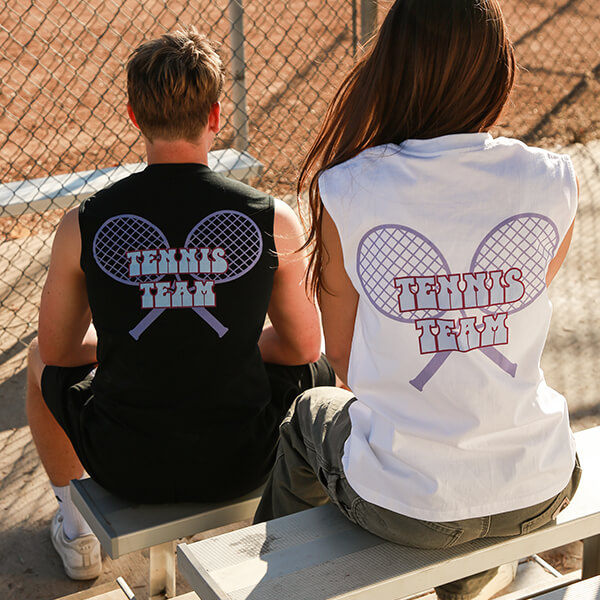 Two people wearing custom tennis team sleeveless shirts sitting on bench by tennis court fence